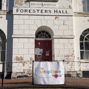 A large doorway on a grand Georgian building with chipped facade. There is a a banner advertising The Scrap Store on railings in front of it.